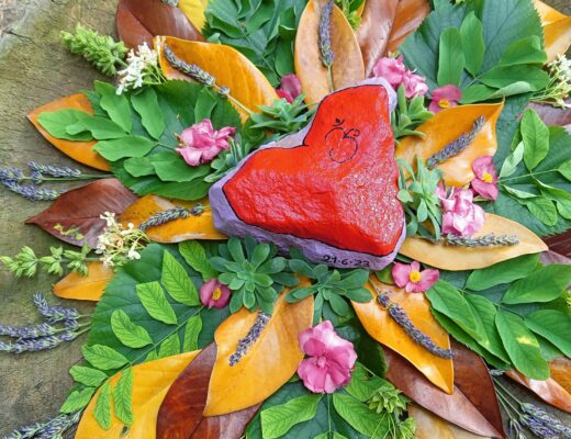 Cuore in pietra dipinto di rosso posto sopra una composizione di foglie e fiori