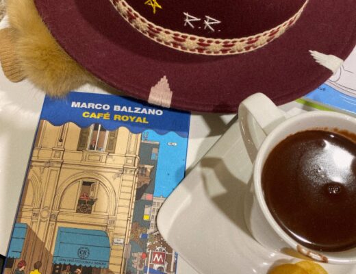 Libro dal titolo Cafè Royal appoggiato accanto a una tazza di cioccolata calda e un cappello